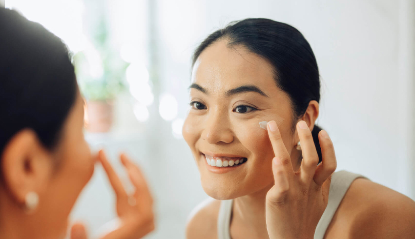 A smiling woman looks at her reflection in a bathroom mirror as she dabs cream with her ring finger onto her upper cheekbone. 
